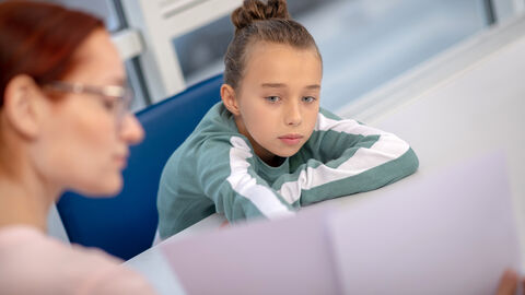 Ein Kind sitzt betrübt an einem Tisch. Eine erwachsene Person sitzt gegenüber und zeigt ihm ein großes Blatt Papier.