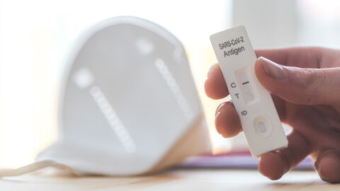 Scharf im Vordergrund ist die Nahaufnahme einer Hand zu sehen, die einen negativen Corona-Selbsttest in den Fingern hält, mit einer FFP2-Maske unscharf im Hintergrund.