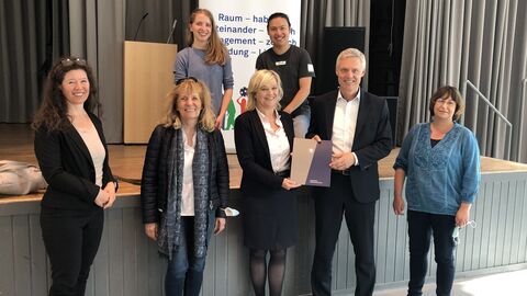 Staatssekretär Lösel mit Beteiligten der Diskussion vor einem Pult und einer Bühne, alle lächeln in die Kamera.