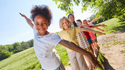 Mehrer Kinder balancieren hintereinander auf einem Ast und lachen in die Kamera. Im Hintergrund Natur.