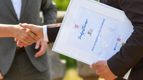 Ein Mann schüttelt die Hand eines anderen. Er hält in der anderen Hand eine Urkunde als "50 Beste Dorfgasthäuser in Hessen" für Hotel Landgasthaus Zum Schäferhof.   