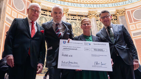 Eine Frau und drei Männer stehen zusammen und blicken freundlich in die Kamera. Zwei Personen halten einen großen Spendenscheck über 10.000 Euro in den Händen.