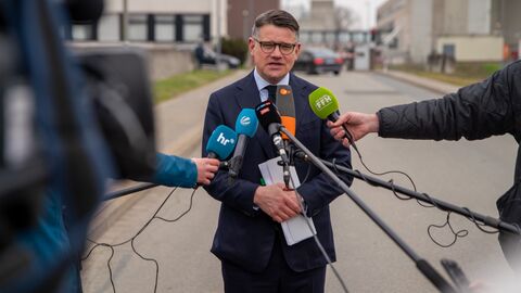 Ministerpräsident Rhein steht in einer Interviewsituation vor Mikrofonen und spricht.