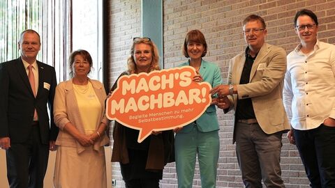 Sechs Personen stehen in einer Reihe - die beiden mittleren halten ein Schild in den Händen mit der Aufschrift "Mach's machbar"