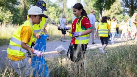 Kinder in Warnwesten sammeln mit Müllzangen Abfall auf einer Wiese an einem Weg.