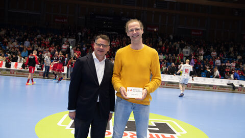 Ministerpräsident Boris Rhein steht auf dem Spielfeld der MT Melsungen. Der Ehrenamtliche Stefan Döring hat von ihm eine Auszeichnung überreicht bekommen. Diese hält er in der Hand.
