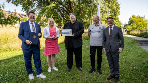 Fünf Menschen stehen nebeneinander auf einer Wiese. Die mittlere Person hält einen Zuwendungsbescheid in der Hand.